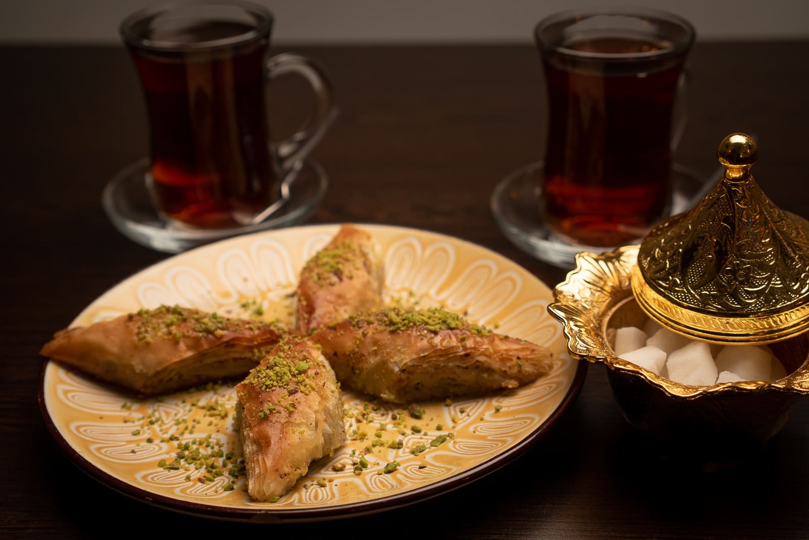 Baklava dusted with pistachio on a patterned plate, beside two glasses of Turkish tea