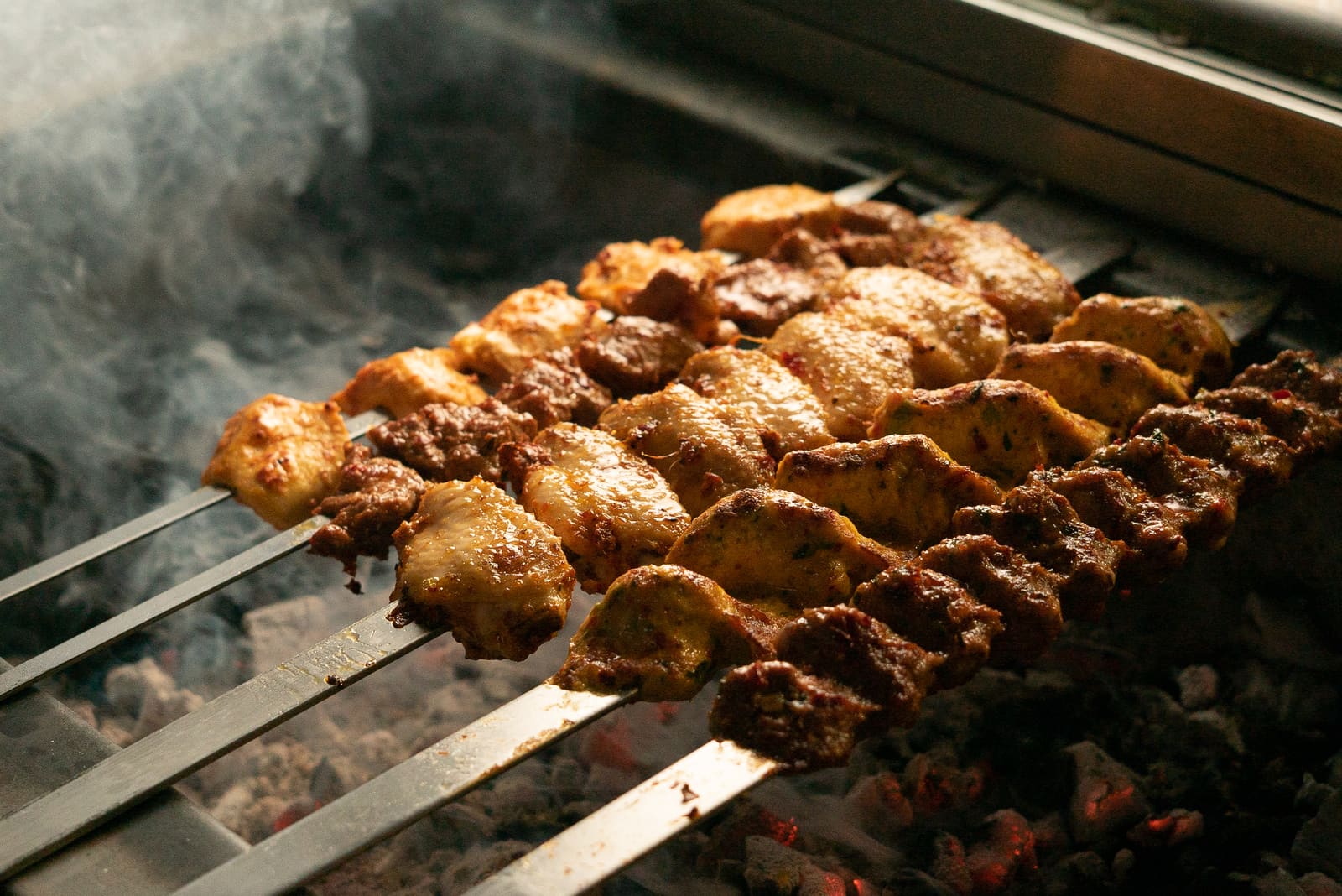 Skewers of chicken and lamb laid across the charcoal grill, smoke rising off the coals